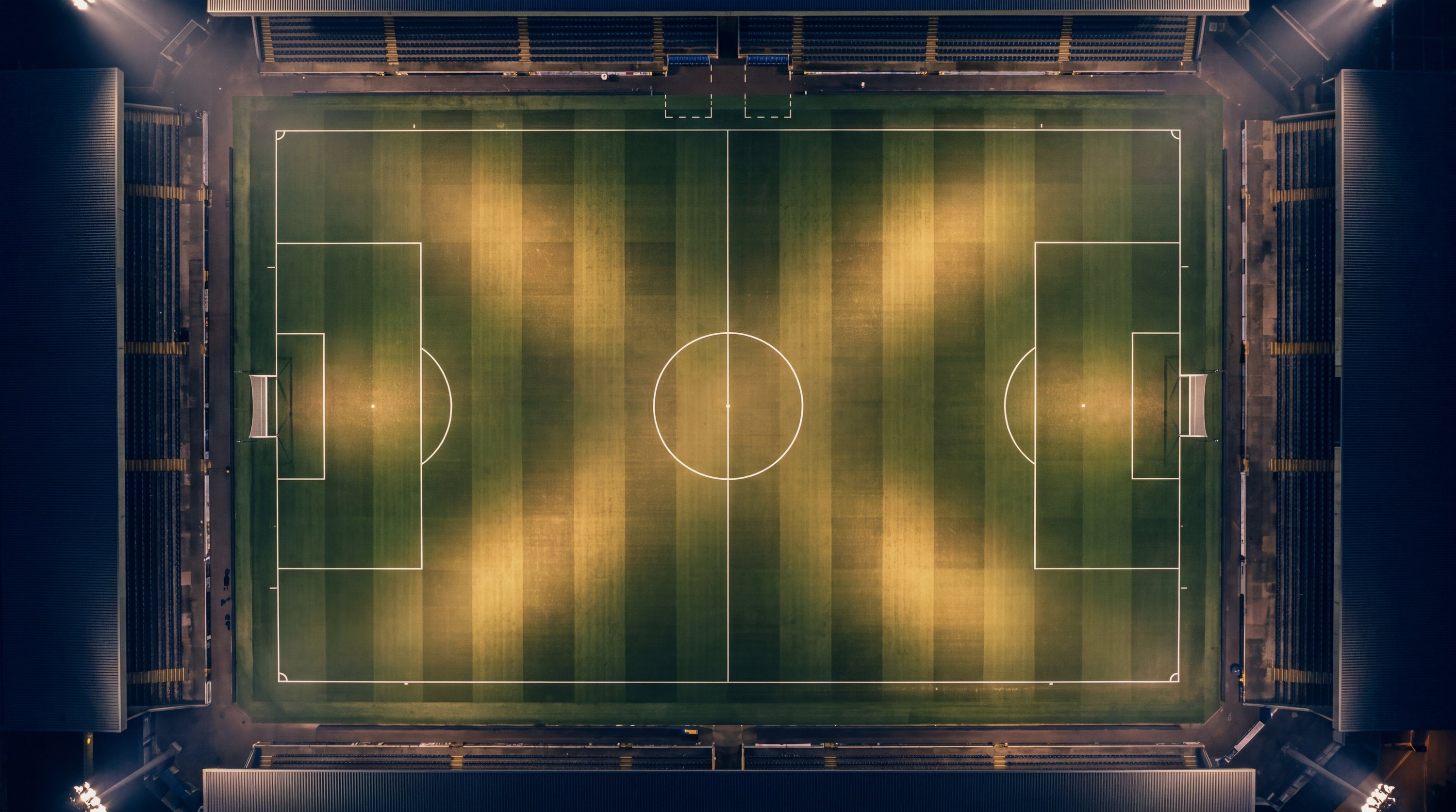 A football pitch from above at night, markings and floodlight pools as clear geometry